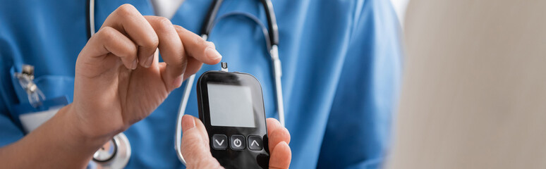 Wall Mural - cropped view of multiracial nurse pointing with finger at glucometer with test strip near senior woman, banner.