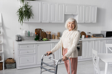 Wall Mural - positive senior woman with grey hair walking with help of walker in modern apartment.