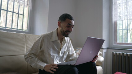 One excited happy young Middle Eastern man reacting positively to successful message online while sitting on couch in front of laptop computer