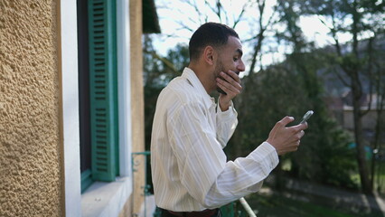 One excited Arab young man reacting with unbelief to positive message on cellphone device while standing outside at balcony during sunny day
