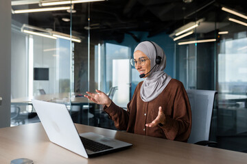 Online teaching. Young Muslim woman in hijab and headset, teacher sitting at desk in office in front of laptop, explaining, talking, gesturing with hands. Conducts distance education.