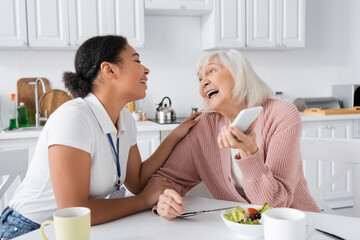 Wall Mural - happy senior woman holding smartphone near cheerful multiracial social worker at home.