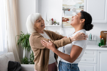 Wall Mural - positive multiracial social worker and senior woman laughing and hugging in living room.