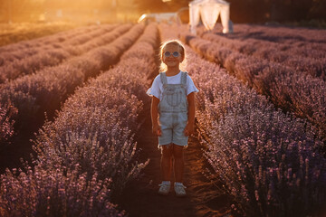 Wall Mural - adorable child girl in lavender field on sunset. smiling kid in sunglasses, jeans jumpsuit is having fun on nature on summer day. Family day, vacation, holiday.International Children's Day,