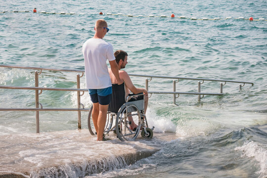 man with disability at beach goes to swimm on a wheelchair with assistance on an accessible ramp.