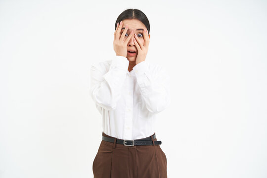 Image of young asian woman with terrified face, hides eyes behind hands and peeks through fingers with scared emotion, white background