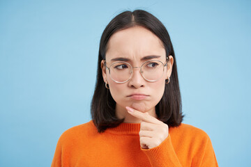 Portrait of thoughtful asian woman in glasses, girl in prescribed eyewear thinking, frowns while looks suspicious, stands over blue background