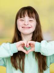Smiling young girl with Downs syndrom shows heart sign at summer park