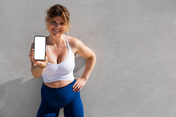 Wall Mural - woman in fitness clothing standing outdoors and showing mobile phone with empty white screen