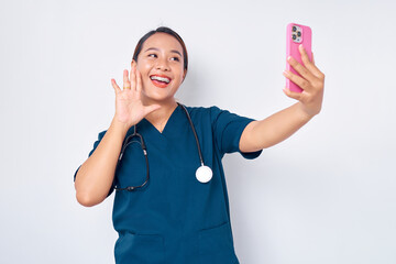 Smiling friendly young Asian woman nurse working wearing blue uniform making video call on smartphone and waving a hand to say hello isolated on white background. Healthcare medicine concept