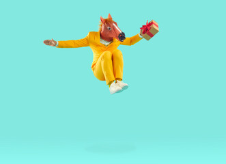 Wall Mural - Full body length shot of happy cheerful joyful excited man in yellow suit and funny silly carnival horse mask jumping up, holding gift present box and flying high in air on blue studio background