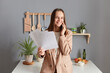 © sementsova321 - Friendly business woman with brown hair wearing beige jacket standing in home kitchen interior talking phone holding papers having conversation with partner.