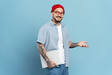 Wall Mural - Authentic portrait of smiling confident asian man, freelancer holding laptop, isolated on blue background. Korean hipster wearing stylish eyeglasses, red hat looking at camera. Successful business