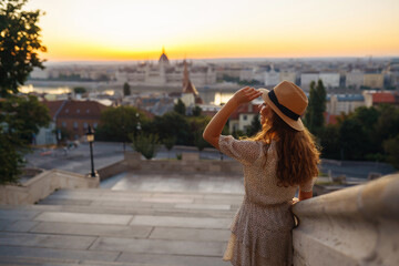 Smiling female tourist in a hat admires the landscape of the city at dawn. Euro-trip. Travel, tourism and active lifestyle concept.