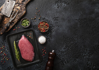 Wall Mural - Raw beef fillet steak in vacuum tray with rosemary on on black background with meat cleavers and pepper grinder.