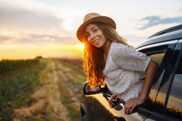 Young happy woman in hat enjoys car ride, leaning out of the window. In summer, a beautiful tourist woman travels by car and enjoys the golden sunset. Concept of an active lifestyle, travel, tourism.