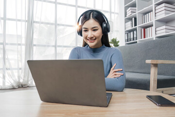 Wall Mural - Smiling young asian woman using laptop web camera while wearing headphones and sitting on the rug beside to the sofa at homes