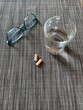 Two capsules on a textured surface, glass of water and a pair of glasses on the side