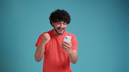 Happy indian man winner using cell mobile phone Excited ethnic guy holding smartphone feels surprised about betting win on cellphone, celebrating success in coral t-shirt on blue studio background