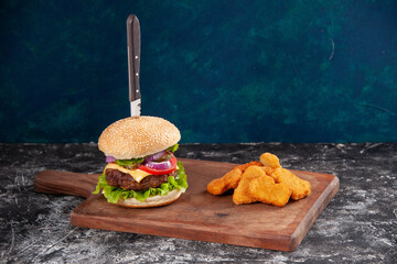 Wall Mural - Knife in tasty meat sandwich and chicken nuggets on wooden cutting board on dark blue background