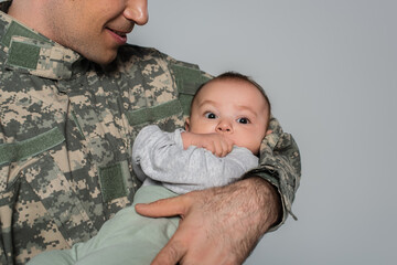 Poster - happy soldier in uniform holding baby boy in arms isolated on grey.