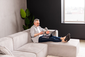 Older man working online with laptop computer at home sitting on couch in living room.