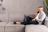 Fototapeta Na ścianę - Happy mature man talking on mobile phone while sitting on a sofa at home with laptop