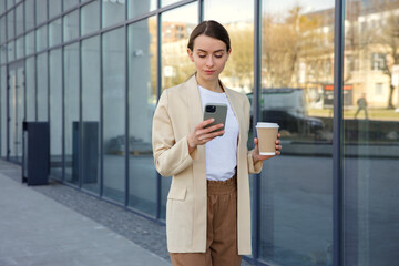 Caucasian business woman walking, using smartphone and holding coffee cup on street. Attractive female typing message near office building during coffee break