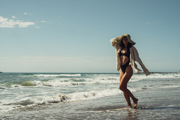 a beautiful girl in a black swimsuit shirt and a straw hat walks on the seashore smiles rejoices laughs