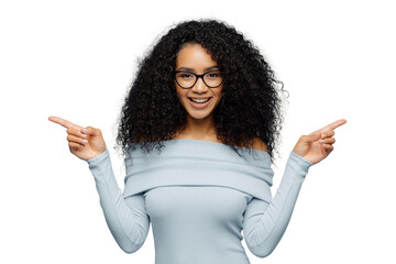 Wall Mural - Happy woman with Afro hairstyle, points sideways with both index fingers, wears casual jumper, hesitates between two objects, isolated over blue background. Female indicates left and right indoor