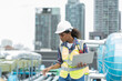 © amorn - Female engineer worker using laptop computer inspecting quality sewer pipes at rooftop of building. African American woman engineer working, checking or maintenance sewer pipes at construction site