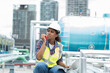© amorn - Female engineer worker using laptop computer inspecting quality sewer pipes at rooftop of building. African American woman engineer working, checking or maintenance sewer pipes at construction site