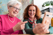 © Studio Marmellata - Positive elderly gray haired mother in pink sweater and eyeglasses pointing at smartphone screen while watching photos with mature redhead daughter at home