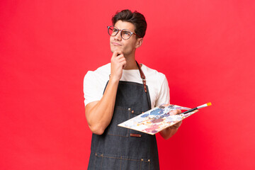 Wall Mural - Young artist caucasian man holding a palette isolated on red background having doubts