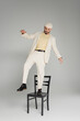 Fashionable homosexual man in suit and beige hat standing on chair on grey background.