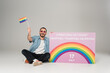 Positive gay man holding lgbt flag near placard with International Day Against Homophobia, Transphobia and Biphobia lettering on grey.