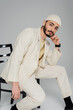 Trendy homosexual man in hat and beige suit posing on chair on grey background.