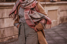 Woman In Autumn Stylish Fashion Brown Long Coat, Scarf And Plaid Pants Walking In The City. Female Casual Street Style Outfit