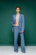 © LIGHTFIELD STUDIOS - full length of stylish woman in satin bra and blue pantsuit posing with hand on hip on turquoise green background.