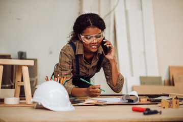 Wall Mural - Happy african american woman using smartphone for confirrm order and customer service, Small business owner young black female carpenter working a wood work in carpentry workshop.