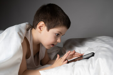 Wall Mural - A 5 year old boy plays games on his phone while lying in bed. Gadgets in bed before bedtime in a child