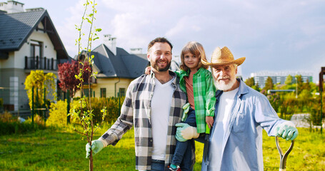 Portrait of happy three male genarations smiling to camera in garden on summer sunny day. Kid planting tree with father and grandfather. Senior man with adult son and little grandson.