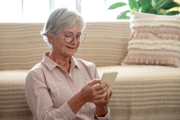 Pretty senior woman relaxed sitting on the floor holding smartphone in hands. Smiling mature lady writing a message, using mobile applications at home.