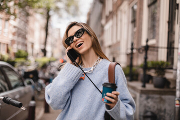 Wall Mural - Beautiful young woman wearing blue sweater, walking outdoors, talking on mobile phone. Beautiful stylish woman talking on phone, calling on mobile phone walking outdoor, hold coffee cup take away.