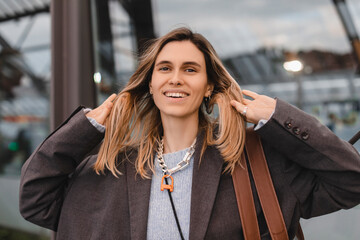 Wall Mural - Attractive happy woman wear gray jacket, blue sweater, brown bag and necklace standing on bus station, fix her hair look flirty. Blonde woman waiting bus look at camera.