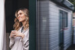 © zvkate - Photo of young curly blonde woman with short hair smiling and looking aside while standing near glass door in house with cup of tea or coffee. Girl with cup of coffee in morning.