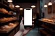 Hand holding phone with white screen and blurred background of a bakery. Concept mockup for app, site, logo... Generative AI