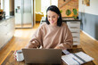© Baba Images - Young adult woman working on her laptop in the kitchen of her home