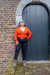 Wall Mural - Latin American senior adult woman standing and leaning against brick wall next to closed wooden door, looking up, long wavy gray hair, wearing low cut orange top, black pants and sunglasses