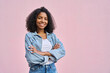 © Stock 4 You - Young stylish woman model pose in denim jacket in front of soft pink background looking at camera. Beautiful curly hair african american girl in trendy clothes with crossed arms smile with copy space.
